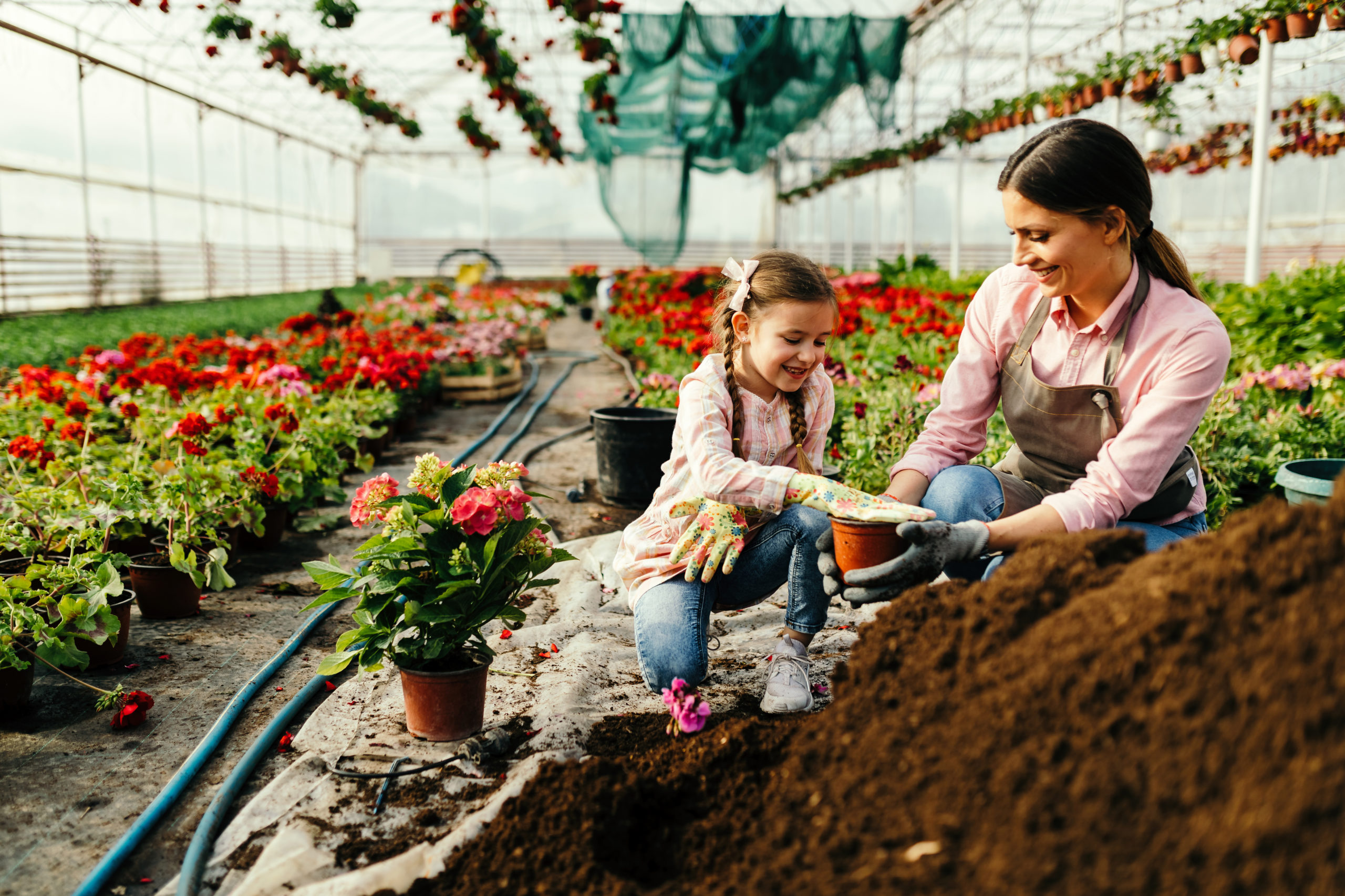 Happy mother and her small daughter working with soil while planting flowers in a greenhouse.