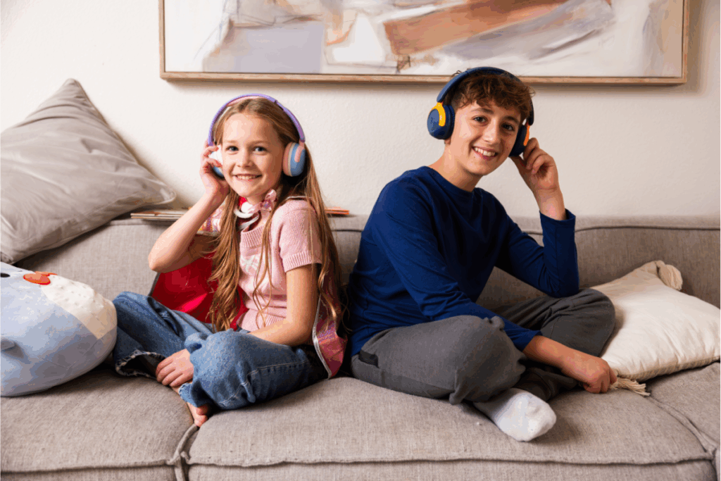 Two kids sitting on a messy couch, both wearing on-ear headphones for children.